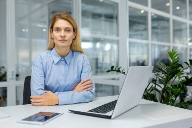 Odaklanmış profesyonel bir kadın ofis masasında oturmuş, ofis malzemeleriyle çevrili modern bir dizüstü bilgisayarla ciddi ciddi çalışıyor..