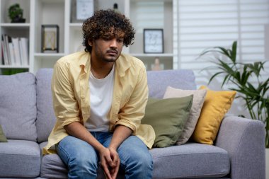 Hintli bir adam, iyi aydınlatılmış bir oturma odasındaki rahat yastıklarla çevrili gri bir kanepede otururken dalgın görünür..