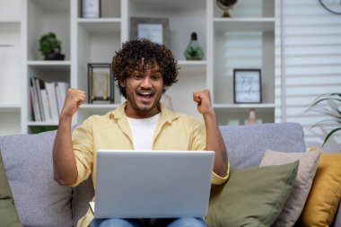 Kanepede oturan, yumruklarını zaferle kaldırmış, dekore edilmiş bir oturma odasındaki dizüstü bilgisayara bakan aşırı mutlu bir birey..