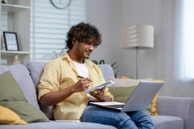 Genç bir yetişkin rahat bir şekilde kanepede oturur, dizüstü bilgisayarla çalışırken, bir not defterine not alır, evden çalışma ortamını betimler..