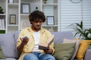 Kanepede oturan kafası karışmış Hintli bir adam elinde telefon ve teslimat kutusuyla internetten olası bir alışveriş dolandırıcılığı için endişeleniyor..