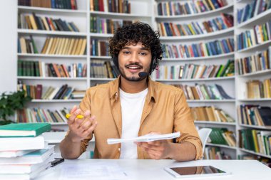 Neşeli genç erkek eğitimci kütüphane ortamında kulaklık ve video çağrı teknolojisi kullanarak çevrimiçi öğretmenlik yapıyor..