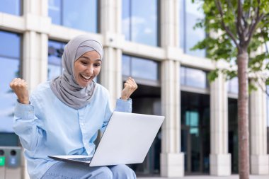 Tesettürlü neşeli bir Müslüman kadın modern ofis binalarının yakınında dizüstü bilgisayar kullanırken başarılı bir şekilde yumruklarını kaldırıyor..