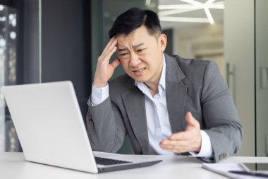 Modern bir ofis ortamında çalışırken bilgisayar ekranında kafası karışık görünen Asyalı bir adam..