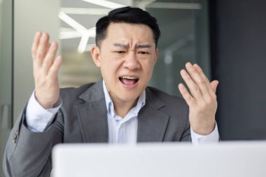 Takım elbiseli sinirli bir Asyalı iş yerindeki stresli bir görüntülü sohbette önünde bir dizüstü bilgisayarla jest yapıyor..