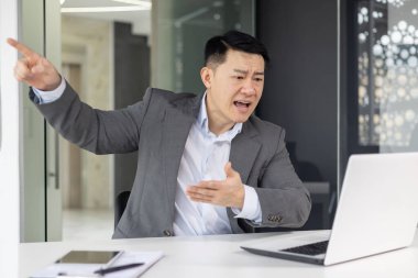 Sinirli Asyalı iş adamı modern ofis ortamında hararetli bir video görüşmesi yaparken el kol hareketi yapıyor..