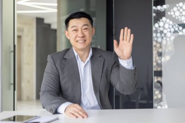 Profesyonel Asyalı iş adamı kameraya bakarken el sallıyor. Modern bir ofisin içinden bir video konferans görüşmesi yapıyor..