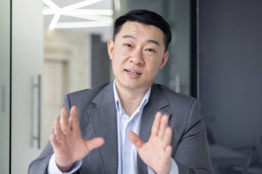 Takım elbiseli profesyonel Asyalı işadamı, modern ofis içinde kameraya dostça bir tavırla hitap ediyor..