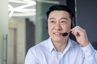 Profesyonel Asyalı iş adamı kulaklık kullanıyor, iyi aydınlatılmış çağdaş ofis ortamında bir sohbete katılıyor..