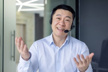 Ofisteki profesyonel Asyalı işadamı kulaklıkla aktif olarak bir video görüşmesine katılıyor..