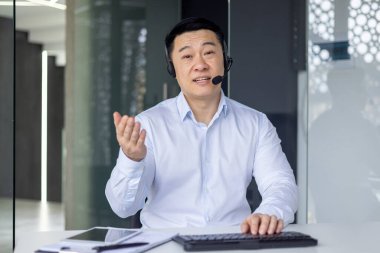 Profesyonel Asyalı bir adam iş ortamında, kulaklıkla arama yapıyor, kameraya bağlanıyor..