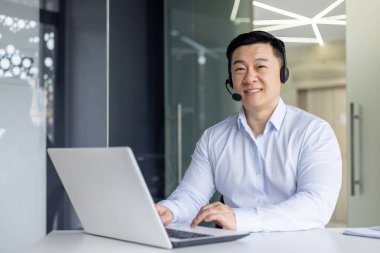 Kulaklıklı neşeli bir Asyalı, modern ofis ortamında dizüstü bilgisayar üzerinde çalışıyor. Profesyonellik ve pozitiflik yayıyor..