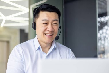 Şirket ofisinde kulaklık ve bilgisayar kullanan Asyalı bir adam ciddi bir ifadeyle işine odaklanmış..