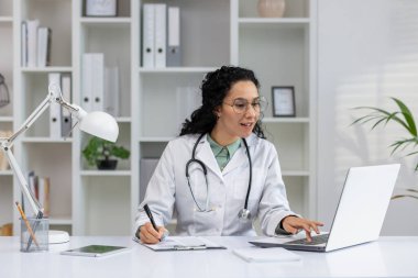 Laboratuvar önlüklü gülümseyen Latin doktor modern bir klinikte bilgisayardaki işiyle meşguldü..