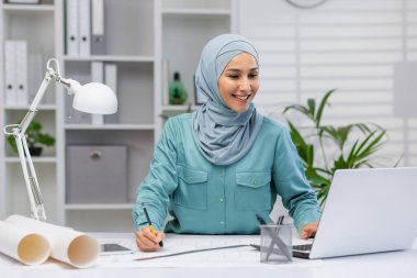 İyi ışıklandırılmış bir ofis alanında planlar ve dizüstü bilgisayarla çalışan gülümseyen Müslüman kadın mimar, odak ve profesyonelliği aktarıyor.