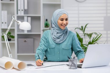 Smiling Muslim female architect working with blueprints and a laptop in a well-lit office space, conveying focus and professionalism. clipart