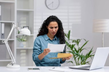 Endişeli Latin iş kadını, modern bir ofis ortamında kötü haberlerle dolu bir belgeyi inceleyerek stres ve hayal kırıklığını dile getiriyor..