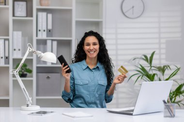 Akıllı telefonu ve kredi kartı olan mutlu İspanyol iş kadını, rahat, modern bir ev ofisinden çalışıyor, özgüvenle gülümsüyor..