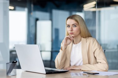 Günümüz ofis ortamında dizüstü bilgisayar üzerinde çalışırken düşünceli bir kadın, kameraya bakıyor..