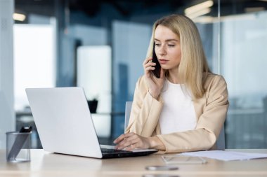 Odaklanmış, bej ceketli bir iş kadını dizüstü bilgisayar kullanıyor ve modern ofis ortamında akıllı bir telefonla konuşuyor..