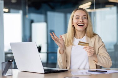 Modern bir ofis ortamında masasında otururken elinde kredi kartı tutan ve mutluluğunu ifade eden neşeli kadın bir profesyonel..