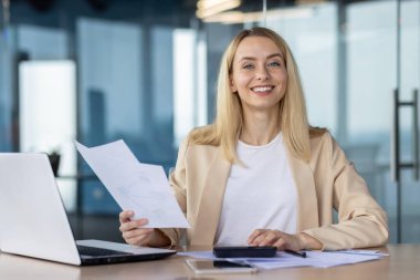 Modern ofis ortamında evraklar ve dizüstü bilgisayarla çalışırken kameraya gülümseyen profesyonel bir kadın yönetici..