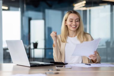 Ofis ortamında neşeli bir iş kadını. Bir belgeye bakarken yumruğunu kaldırıp seviniyor..