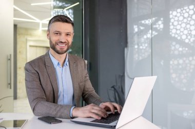 Günümüz ofis ortamında dizüstü bilgisayarı üzerinde çalışırken kameraya bakan kendine güvenen bir erkek profesyonel..