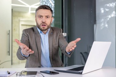 Ofis ortamında kafası karışmış bir işadamının portresi. Kafası karışmış bir ifadeyle doğrudan kameraya bakıyor ve sorgulayıcı bir şekilde ellerini kaldırıyor..