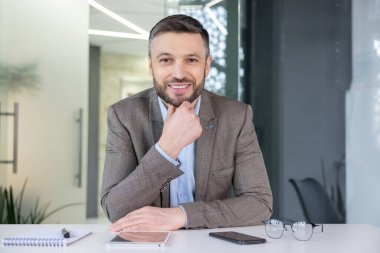 Kendine güvenen profesyonel bir erkek kameraya bakıyor, modern ofis ortamında dostça bir gülümsemeyle kameraya bakıyor..