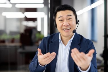 Dost canlısı Asyalı iş adamı kulaklık takıyor ve modern ofis ortamında müşterilerle veya takım üyeleriyle iletişim kuruyor..