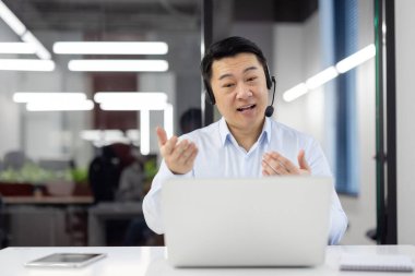 Neşeli Asyalı iş adamı bir ofis ortamında dizüstü bilgisayar ve kulaklıkla görüntülü konuşma yapıyor..