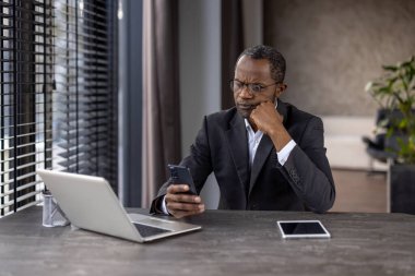 Modern bir ofis masasında akıllı telefon ve dizüstü bilgisayarla çalışan takım elbiseli ciddi bir işadamı..