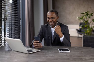 A smiling African businessman celebrates success while using his smartphone and laptop in a modern office setting. clipart