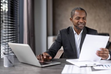 Takım elbiseli iş adamı laptopla çalışıyor ve modern ofis masasında kağıt dökümanlarını inceliyor..