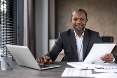 Profesyonel Afro-Amerikan işadamı, modern bir ofis ortamında, masasında dizüstü bilgisayar ve tabletle evrakları analiz ediyor..