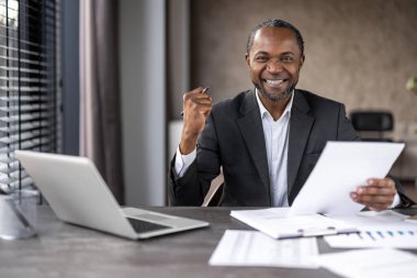 Başarılı bir Afrikalı Amerikalı işadamı, ofis masasında dizüstü bilgisayar ve evrak işlerini incelerken gülümsüyor..