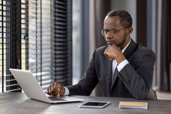 A professional businessman in a suit concentrating on his work on a laptop at a modern office desk, possibly analyzing data or planning strategies.
