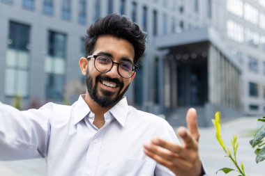Gözlüklü, gülümseyen bir Hintlinin yakın plan fotoğrafı. Ofis merkezinin yanında duran beyaz tişörtlü ve elinde tuttuğu bir telefonun kamera önünde konuşan..