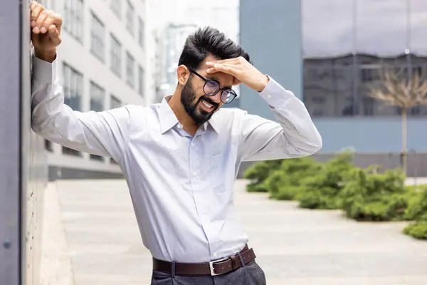 Tired and exhausted Indian young businessman standing near working offices outside and holding his hand, feeling pain and dizzy.