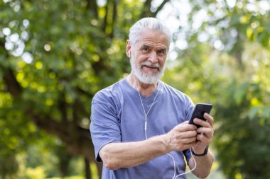 Parkta duran, kulaklık takan, telefon tutan, spor yapan, yürüyen ve kameraya gülümseyen yaşlı, gri saçlı bir adamın yakın plan portresi..