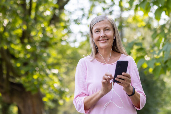 Close-up portrait of a smiling gray-haired sporty woman standing in the park wearing headphones, holding a phone and looking at the camera.