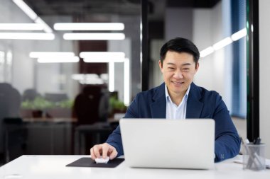 Başarılı ve gülümseyen Asyalı takım elbiseli genç işadamı modern ofiste oturuyor ve dizüstü bilgisayarla çalışıyor..