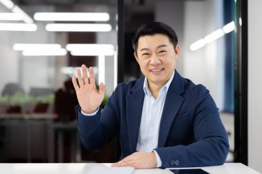 Gülümseyen ve başarılı Asyalı iş adamı, ofis masasında oturuyor, kameraya bakıyor, selamlıyor ve el sallıyor..