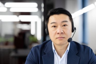 Ofiste oturan ve kameraya güvenle bakan, kulaklık takan ciddi bir Asyalı adamın yakın plan portresi..