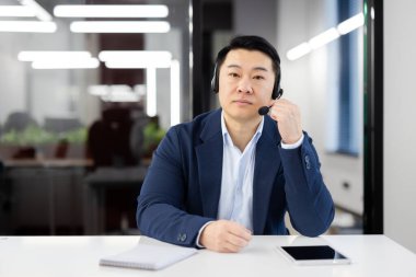 Ofiste çalışan ciddi ve kendinden emin genç bir Asyalı 'nın portresi. Masada oturmuş kameraya bakıyor..