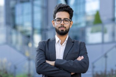 Sokakta takım elbiseli, gülümseyen ve başarılı genç Hintli bir işadamının portresi. Kollarını göğsünün üzerinden geçirip kendine güvenle kameraya bakıyor..