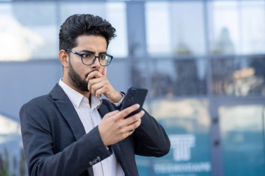 Endişeli genç Hintli takım elbiseli adam ofis binasının dışında duruyor cep telefonu ekranına bakıyor, ağzı elle kapatıyor, kötü haberleri okuyor..
