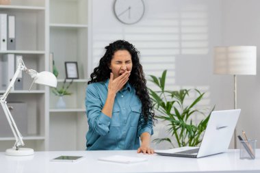 Çok çalışan kıvırcık saçlı iş kadını kendini yorgun hissediyor. Modern ofis alanında dizüstü bilgisayar ve ofis malzemeleriyle esniyor..
