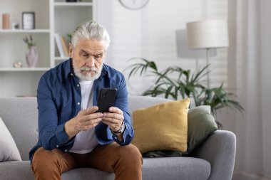 Gri saçlı olgun bir adam modern oturma odası ortamında akıllı telefonuna yoğunlaşarak yaşlılar arasında teknoloji kullanımı kavramını aktarıyor..